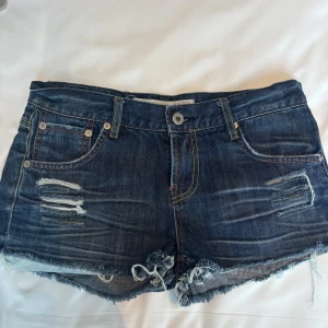 Blå jeansshorts från Youdally - Snygga blå jeansshorts från Youdally med slitna detaljer och fransig kant. Klassisk femficksmodell med gul kontrastsöm och coola broderier på bakfickorna. Perfekt för dig som gillar en avslappnad och trendig stil.