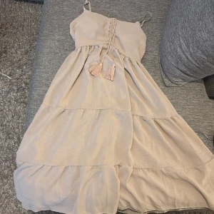 Beige långklänning med snörning - Säljer en beige långklänning med tunna axelband och snygg snörning framtill med tofsar. Klänningen har volanger och ett luftigt, flowy tyg som ger en chill vibe. Perfekt för varma dagar och har en romantisk känsla med sin ljusa ton.