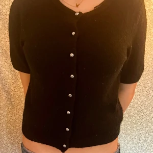 Svart stickad topp med knappar - Svart stickad topp med korta ärmar och rund halsringning. Toppen har en rad silvriga knappar framtill som ger en snygg detalj. Använd 1 gång.