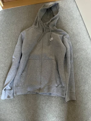 Grå hoodie från Nike med dragkedja - Grå hoodie från Nike med vit broderad logga på bröstet. Tröjan har huva med snörning, två fickor fram och hel dragkedja. Mjuk insida i fleece och långärmad passform. Perfekt för chill eller träning.