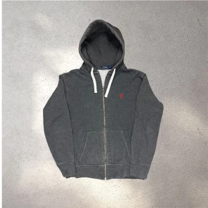 Mörkgrå hoodie från Polo Ralph Lauren - Säljer en mörkgrå hoodie från Polo Ralph Lauren med dragkedja och vit snörning i huvan. Mycket bra skick (dock något tvättpåverkad, lite hårdare än vanligt) skickar snabbt efter att du har beställt✅📦