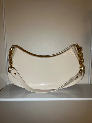 Beige axelväska från Primark med kedja - Stilren beige axelväska från Primark i glansigt syntetmaterial. Väskan har en snygg bågformad design, guldfärgade kedjedetaljer och dragkedja upptill. Perfekt storlek för det viktigaste och ger en trendig touch till din outfit. Observera lite sliten men i god skick! 