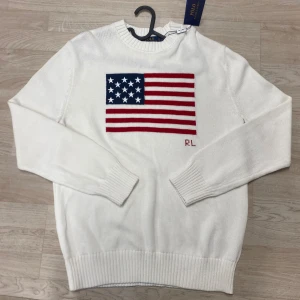 Vit stickad tröja Polo Ralph Lauren - Vit stickad tröja från Polo Ralph Lauren med amerikansk flagga broderad på bröstet och RL-detalj i rött. Tröjan har rund hals, ribbade muddar och lång ärm. Perfekt för dig som gillar klassisk och clean stil med en cool twist.