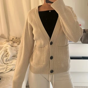 Beige stickad kofta med knappar - Säljer en beige stickad kofta med v-ringad hals och tre svarta knappar framtill. Koftan har två praktiska fickor och ribbade muddar vid ärmslut och nederkant. Perfekt för lager-på-lager och chill stil.