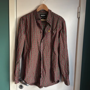 Rutig röd skjorta från Lyle & Scott - Snygg rutig skjorta från Lyle & Scott i slim fit med klassisk krage och långa ärmar. Färgerna är röd, grön, vit och svart, och materialet är mjuk bomull. Bröstficka med gul broderad logga och svarta knappar på framsidan och vid manschetterna. Perfekt för dig som gillar stilrena detaljer.