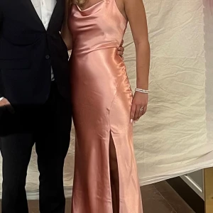 Ljusrosa långklänning med slits - Säljer en elegant ljusrosa långklänning i satin med smala axelband och öppen rygg med knyt. Klänningen har en snygg slits framtill och ett mjukt, glansigt material som ger en lyxig känsla. Perfekt för bal eller fest! Passformen är slim och klänningen har en draperad urringning. (Klänningen har några små fläckar allra längst ner på släpet men det syns inte alls när man har klänningen på sig!!) 