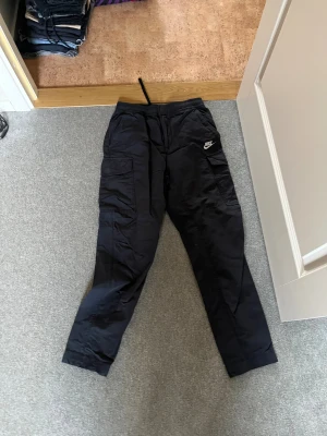 Svarta cargopants från Nike - Svarta cargopants från Nike med broderad logga på benet. Byxorna har flera fickor, elastisk midja med snörning och är tillverkade i ett slitstarkt syntetmaterial. Perfekta för en chill streetwear-look.
