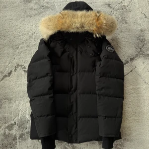 Canada Goose Wyndham parka - Black label - Svart Canada Goose-dunjacka med stor huva och avtagbar beige/brun päls. Ribbade muddar, knappar och dragkedja framtill samt patch med logga på ärmen. Varm och snygg puffer-modell, perfekt för vintern.  Skonsamt använd och utan synliga skador. Köpt för några år sedan men passar inte längre då jag vuxit ur den. Tags finns kvar, men kvitto och box saknas eftersom jag förlorat min gamla mail. Autentisering kan visas.  Pris: 6 499 kr