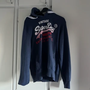 Mörkblå Superdry hoodie med tryck - Säljer en mörkblå hoodie från Superdry med stort vitt och rött tryck på bröstet. Tröjan har huva, känguruficka och långa ärmar. Perfekt för dig som gillar streetwear och vill ha en snygg och bekväm tröja med cool logga.