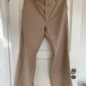 Beiga kostymbyxor från Stockholm Surfboard Club - Snygga beiga kostymbyxor från Stockholm Surfboard Club i klassisk rak modell med vida ben. Byxorna har diskreta fickor bak och dold dragkedja fram. Perfekt för dig som gillar stilrena och trendiga plagg med lite edge.