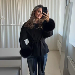 Gina tricot jacka  med fuskpälsärm - Svart omlottjacka från Gina Tricot med knytband i midjan och breda ärmar med fluffiga fuskpälsdetaljer vid ärmsluten. Jackan har en loose fit och är perfekt för dig som gillar en trendig och chic look. Enkel, stilren och riktigt snygg. Jag har köpt storlek L pga att den är från kids avdelningen. Skulle säga att den sitter som en S på mig som vanligtvis bär xs/s o är 173