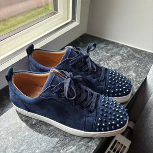 Christian Louboutin mörkblåa - Snygga mörkblå sneakers i mocka från Christian Louboutin med ikoniska röda sulor och coola nitar på tån. Skorna har snörning och en vit platt sula. Perfekta för dig som vill sticka ut med exklusiv streetstyle. Oanvända hör av er vid frågor.