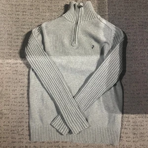 Grå stickad half zip Peak Performance - Snygg grå stickad tröja från Peak Performance med half zip och hög krage. Tröjan har ribbade muddar och ärmar, samt en diskret svart logga broderad på bröstet. Perfekt för dig som gillar stilren och sportig look. Passar dig som är 175 typ  och väger 65kg