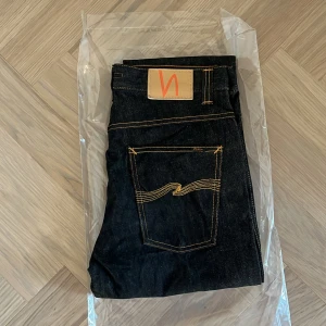 Helt nya Nudie jeans - slim Jim - W32 L32 - Helt nya och oanvända Nudie Jeans Co Slim Jim i den klassiska tvätten Dry Streaky – fortfarande i originalförpackning (se bilder).  Detta är en tidlös modell från Nudie Jeans med rak passform, perfekt balans mellan klassiskt snitt och modern stil, tillverkade i högkvalitativ japansk denim.  Detaljer: • Modell: Slim Jim • Storlek: W32 / L32 • Färg: Dry Streaky (mörk indigo med subtilt randig struktur i tyget) • Passform: Regular slim – normal midja, lätt avsmalnande ben