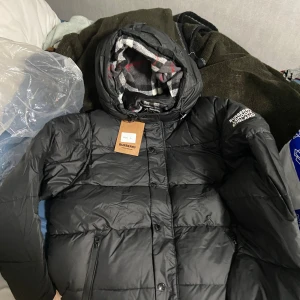 Svart pufferjacka från Burberry L - Svart pufferjacka från Burberry med klassisk rutig huva i grått, vitt och rött. Jackan har dragkedja, knappar och två sidofickor. Perfekt för kalla dagar med sin tjocka och quiltade design. Stort Burberry-tryck på axeln och snygga detaljer.