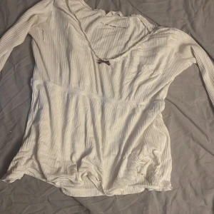 Vit ribbad långärmad tröja med rosett - Vit långärmad tröja med ribbad struktur och v-ringad hals. Söt liten rosett framtill vid halsringningen. Materialet känns mjukt och lätt, perfekt för dig som gillar en enkel men gullig stil. Passar dig som gillar detaljer och vill ha något som sticker ut lite. Har ett hål vid sidan av bröstet som man kan sy ihop