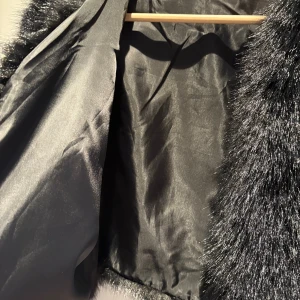 Svart croppad pälsjacka - Snygg svart croppad pälsjacka med långärmade ärmar och glansig insida. Jackan har en fluffig och lyxig look som verkligen sticker ut. Perfekt för dig som vill ha en statement piece i garderoben. Jackan är i syntetiskt pälsmaterial och har en stilren design utan synliga knappar eller dragkedja.