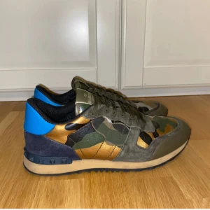 Valentino Rockrunner sneakers camouflage - Säljer ett par Valentino Rockrunner sneakers med grymt camouflage-mönster i grönt, svart, guld och blått. Skorna har detaljer i mocka och skinn, platt sula med nitar och snörning. Perfekt för dig som vill sticka ut med lyxiga och unika sneakers.