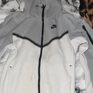 Nike grå och vit vindjacka med huva - Nike vindjacka i ljusgrått och vitt med svart logga på bröstet. Jackan har hel dragkedja, huva och snygga svarta detaljer längs bröstet. Materialet är mjukt och känns sportigt, perfekt för chill eller utomhus. Långärmad och med stilren design.