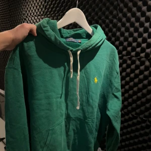 Grön hoodie från Polo Ralph Lauren - Snygg grön hoodie från Polo Ralph Lauren med klassisk gul logga på bröstet. Tröjan har huva med vit snörning, känguruficka och ribbade muddar. Perfekt för chill dagar och enkel att matcha med jeans eller joggers.