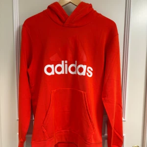 Röd Adidas hoodie med huva - Säljer en röd hoodie från Adidas med stor vit logga på bröstet. Tröjan har huva, lång ärm och en stor magficka framtill. Perfekt för chill dagar eller när du vill ha en sportig look. Storken är M och skicket 9/10