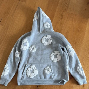 Grå hoodie med vita Denim Tears-märken - Säljer en grå hoodie från Denim Tears med unika vita applikationer i form av blommor över hela tröjan. Tröjan har huva, magficka och ribbade muddar. Perfekt för dig som gillar streetwear och vill sticka ut med cool design.