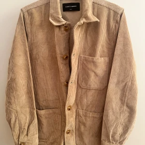 Beige manchester overshirt från A Day's March - Säljer en beige overshirt från A Day's March i mjuk manchester. Jackan har klassisk krage, tre stora fickor framtill och stängs med knappar. Perfekt för lager-på-lager och har en chill, loose passform som funkar till många stilar. Jackan är i 100% bomull.