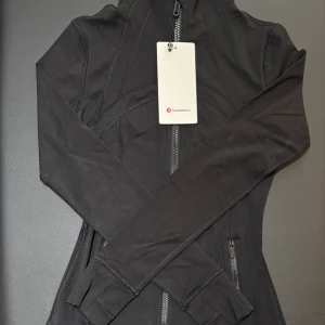 Svart Define jacka från Lululemon storlek 4 - Svart, slim fit Define jacka från Lululemon storlek 4 med dragkedja, zip-fickor och tumhål. Tillverkad i mjukt Nulu-material med Lycra för stretch och form. Jackan har en ventilerande ryggdetalj och sitter snyggt på höften. Perfekt för träning och aktiva dagar.