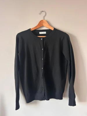 Svart kofta från Primark Cares - En stilren svart kofta från Primark Cares i storlek M. Koftan har rund hals, långa ärmar och knäppning med svarta knappar framtill. Materialet är mjukt och känns skönt mot huden, perfekt för lager-på-lager. Enkel och klassisk design som funkar till många outfits.