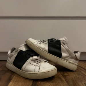 Valentino Open sneakers vit/svart (äkta) - Snygga Valentino Open sneakers i vitt skinn med bred svart läderpanel över snörningen. Platt sula och rund tå, samt ikoniska nitar på hälen. Perfekt för dig som gillar lyxiga streetwear-vibbar och vill sticka ut med designer på fötterna.