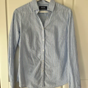 Blårandig skjorta från Sebago - Snygg blårandig skjorta från Sebago i storlek Small. Skjortan har klassisk krage, vita knappar framtill och långa ärmar med manschett. Tillverkad i mjuk bomull som känns skön mot huden. Perfekt för dig som gillar stilrena och fräscha plagg.