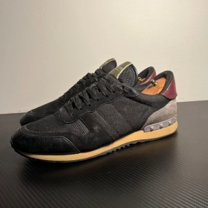 Valentino Garavani Rockrunner sneakers - Svarta Valentino Garavani Rockrunner sneakers med detaljer i mocka, skinn och mesh. Snygg kontrast med beige sula och vinröd häl. Ikoniska nitar på bakkappan och grönt logomärke på plösen. Rund tå, snörning och platt sula för en cool och sportig vibe.