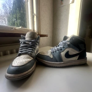 Nike Air Jordan 1 Mid blå/vit - Nike Air Jordan 1 Mid sneakers i blått och vitt med klassisk snörning och rund tå. Skorna har en snygg kontrast mellan ljusblå och vit skinnpanel, samt den ikoniska swoosh-loggan på sidan. Perfekt för dig som gillar streetwear och vill sticka ut.