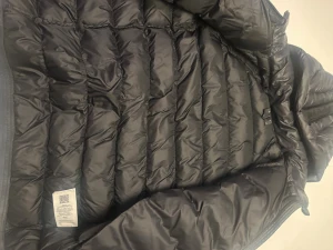 Svart dunjacka Peak Performance - Svart dunjacka från Peak Performance med huva och broderad logga på bröstet och huvan. Jackan har quiltad design, dragkedja framtill och sidofickor med dragkedja. Perfekt för kalla dagar och har en clean, sportig look. Kostar 2000 ny