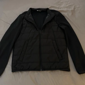 Svart quiltad pufferjacka från Zara - Svart pufferjacka från Zara med quiltad framsida och släta ärmar. Jackan har två snedställda fickor, hel dragkedja och en enkel ståkrage. Materialet är syntet och polyester, perfekt för kyliga dagar. Stilren och enkel design som funkar till det mesta.