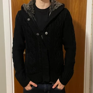 Svart stickad cardigan med fuskpäls - Svart stickad cardigan med grov kabelstickning och mysig huva fodrad med gråmelerad fuskpäls. Jackan har dragkedja och knappar framtill, ribbade muddar och en loose passform. Perfekt för kalla dagar när du vill vara både varm och chill.