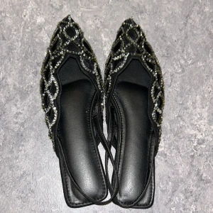 Svarta ballerinaskor med glittriga detaljer - Snygga svarta ballerinaskor med spetsig tå och glittriga pärldetaljer i silver och guld. Skorna har ett nätliknande mönster och platt sula, vilket ger en cool och elegant vibe. Perfekta för dig som gillar att sticka ut med dina skor.
