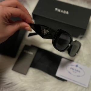 Svarta solglasögon från Prada - Snygga svarta solglasögon från Prada med fyrkantig form och breda skalmar. Modellen är SPR17NS och har en stilren, modern look med diskret logga på sidan. Tillverkade i blankt syntetmaterial och levereras med originalbox, fodral och certifikat.