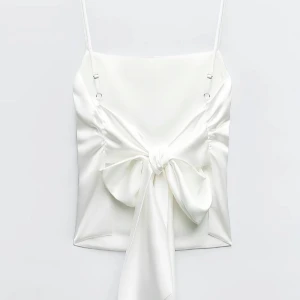 Svart satintopp med rosett - INTRESSEKOLL - Superfin vit linnetopp i satin med smala axelband och stor rosett framtill. Toppen har en slät baksida och snygga rynkade detaljer vid sidorna. Perfekt för dig som vill sticka ut med en elegant och trendig look.