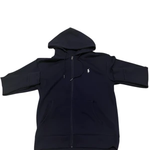 Marinblå hoodie från Polo Ralph Lauren - Snygg marinblå hoodie från Polo Ralph Lauren med dragkedja och huva. Tröjan har det klassiska broderade Polo-logot på bröstet, två fickor framtill och ribbade muddar vid ärmslut. Perfekt för chill dagar och enkel att matcha med jeans eller joggers.