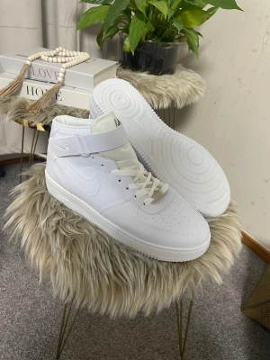 Nike Air Force 1  - Nike Air Force 1 Highs i helvit färg med klassisk snörning och kardborreband vid ankeln. Skorna har en platt sula med cirkulärt mönster, perforerad tå och rund form. Tillverkade i syntetmaterial och skinn för en clean och fräsch look.