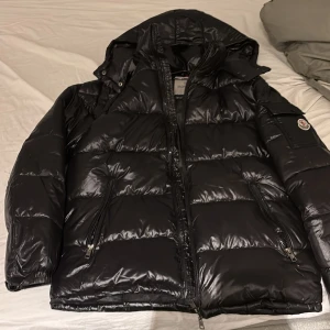 Svart Moncler dunjacka med huva - Svart dunjacka från Moncler med glansig finish, huva och dragkedja framtill. Jackan har två sidofickor med dragkedja och en ficka med Moncler-logga på ärmen. Fylld med vitt gåsdun och har coolt serietryck på insidan.