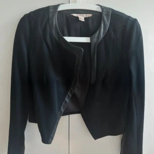 Svart kavaj jacka från Diane von Furstenberg - Snygg svart kavaj från Diane von Furstenberg med coola skinn-detaljer vid krage och ärmslut. Croppad passform och långärmad design, perfekt för dig som gillar stilrena och moderna plagg. Kavajen har knappar på ärmarna och en öppen front. Tyvärr har jackan smink defekt vid etiketten vid nacken. Jackan är i storlek US 2