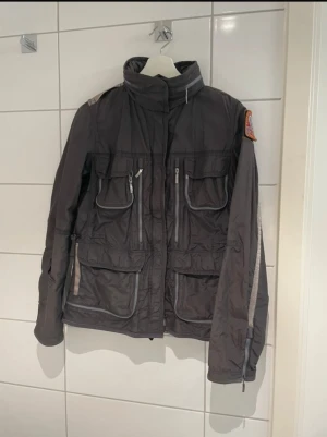Mörkgrå vindjacka Parajumpers M - Snygg mörkgrå vindjacka från Parajumpers med coolt patch på ärmen, flera fickor med dragkedja och hög krage. Jackan har detaljer i grått och orange, meshfoder och sportig look. Perfekt för dig som gillar streetstyle och funktion.