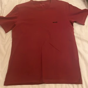 Vinröd t-shirt från BOSS, storlek M - Snygg vinröd t-shirt från BOSS med diskret logga på bröstet. Klassisk rund hals och korta ärmar. Tillverkad i mjuk bomull som känns skön mot huden. Perfekt för dig som gillar stilrena och enkla plagg med premiumkänsla.