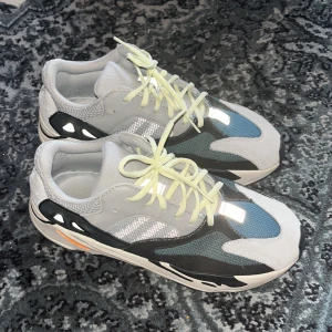 Adidas Yeezy Boost 700 Wave Runner - Säljer ett par Adidas Yeezy Boost 700 Wave Runner sneakers med chunky siluett. Skorna har en mix av grått, svart, blått och orange detaljer, mesh och mocka på ovandelen samt reflekterande inslag. Platt sula med unik mönstring och snörning i ljusgul färg.
