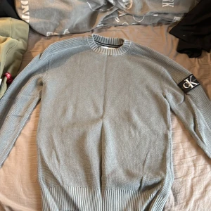 Ljusgrå stickad tröja Calvin Klein Jeans - Snygg ljusgrå stickad tröja från Calvin Klein Jeans med ribbad krage och muddar. Tröjan har långärmad passform och en stor CK-logga på vänster ärm som ger en cool vibe. Perfekt för dig som gillar stilrena och moderna plagg.