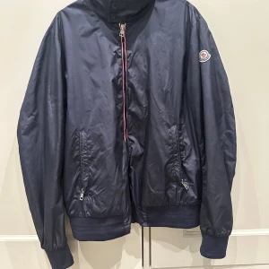 Mörkblå vindjacka från Moncler - Snygg mörkblå vindjacka från Moncler med klassisk logga på ärmen. Jackan har dragkedja med röd och vit detalj, två fickor med dragkedja och ribbade muddar vid ärmslut och nederkant. Perfekt för dig som gillar stilren och sportig look.