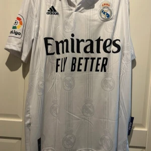 Real Madrid Benzema matchtröja Adidas - Säljer en vit Real Madrid fotbollströja från Adidas med Benzema och nummer 9 på ryggen. Tröjan har korta ärmar, La Liga-märke på ärmen och diskret klubbmönster i tyget. Materialet är lätt och andas, perfekt för fotboll eller supporterliv.