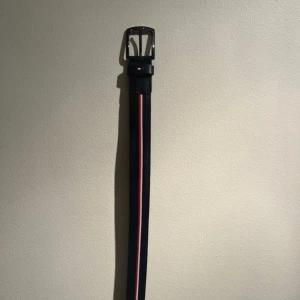 Svart randigt bälte Tommy Hilfiger - Snyggt svart bälte från Tommy Hilfiger med en randig detalj i rött och vitt längs hela bältet. Klassisk metallspänne och diskret logga vid fästet. Perfekt accessoar för att lyfta din outfit med en sportig touch.